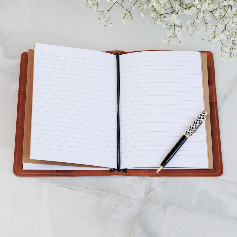 May include: An open, lined notebook with a brown leather cover and a black elastic closure. A black and gold pen with a patterned design rests on the right page. White flowers are in the background.