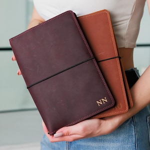 May include: Two leather journals in burgundy and tan, held together with a black elastic band. The burgundy journal has gold initials "NN" embossed on the cover. The tan journal has gold initials "AG".