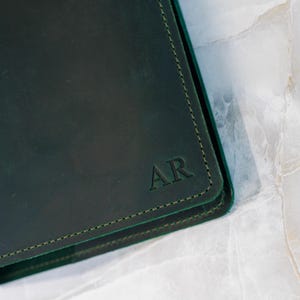 Pu&ograve; includere: Quaderno in pelle verde scuro con le iniziali "AR" in rilievo nell'angolo. Il quaderno ha un bordo cucito in un colore verde pi&ugrave; chiaro. La pelle ha una leggera lucentezza.
