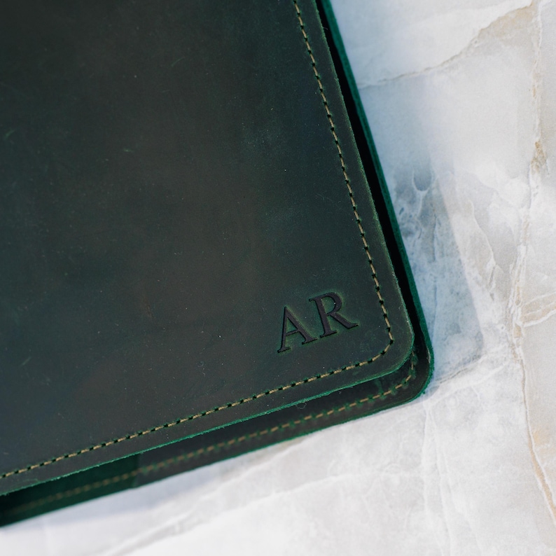 Pu&ograve; includere: Quaderno in pelle verde scuro con le iniziali "AR" in rilievo nell'angolo. Il quaderno ha un bordo cucito in un colore verde pi&ugrave; chiaro. La pelle ha una leggera lucentezza.