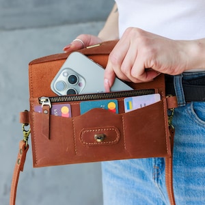 Puede incluir: Bolso bandolera de cuero marrón con múltiples compartimentos, incluyendo un bolsillo con cremallera y ranuras para tarjetas. Un smartphone está colocado dentro del bolso. El bolso tiene una correa larga y un cierre de broche.