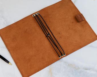Op de afbeelding: Open, bruin leren notitieboek met zwarte elastische sluitingen en een leren lipsluiting. Het notitieboek ligt open op een wit marmeren oppervlak, met een zwarte pen in de hoek.