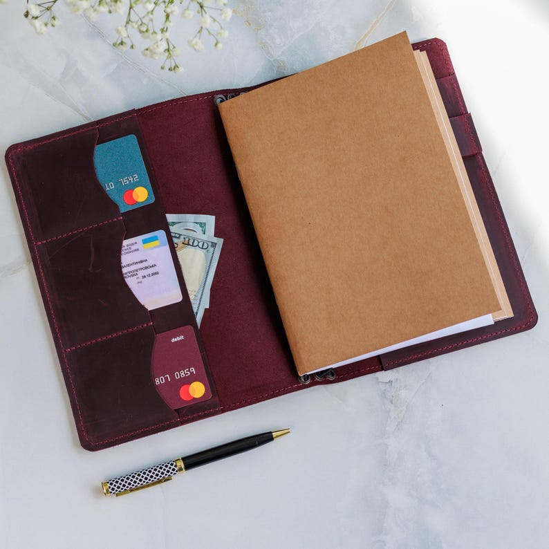 May include: Open burgundy leather journal with card slots and a brown notebook. The journal contains credit cards and cash. A black and white patterned pen with a gold tip rests beside the journal.