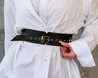 May include: A black leather belt with gold-coloured metal hardware. The belt has a wide design and features a buckle closure. The belt is worn over a white button-down shirt.