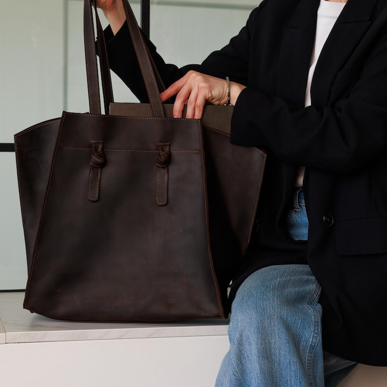 May include: A dark brown leather tote bag with a rectangular shape and two handles. The bag has a front pocket and decorative knots. The bag is being held by a person wearing a black blazer and blue jeans.