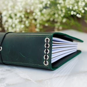 Pu&ograve; includere: Un diario in pelle verde scuro con una chiusura elastica nera. Il dorso del diario &egrave; cucito con occhielli argentati e filo nero. Le pagine sono bianche e la copertina &egrave; leggermente aperta.