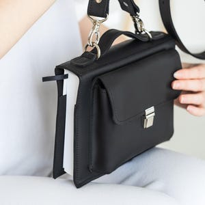Peut inclure: Un sacoche en cuir noir avec une poche à rabat et des accessoires argentés. Le sac a une poignée supérieure et une bandoulière amovible. Un signet noir est visible sur le côté. Le sac est conçu pour contenir un livre.