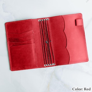Pu&ograve; includere: Vista dall'alto di un'agenda in pelle rossa aperta. Presenta fessure per carte a sinistra e un bordo smerlato decorativo a destra. Anelli metallici al centro per contenere le pagine. Il testo "Color: Red" si trova nell'angolo in basso a destra.
