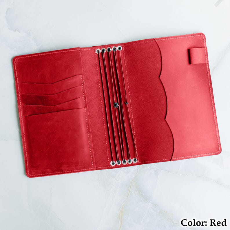 Pu&ograve; includere: Vista dall'alto di un'agenda in pelle rossa aperta. Presenta fessure per carte a sinistra e un bordo smerlato decorativo a destra. Anelli metallici al centro per contenere le pagine. Il testo "Color: Red" si trova nell'angolo in basso a destra.