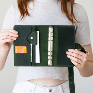 Puede incluir: Una agenda de cuero verde oscuro con seis anillas, que incluye un bolsillo interior con cierre de botón a presión, una ranura vertical para tarjetas con una tarjeta de crédito naranja y un mecanismo de anillas de metal plateado.