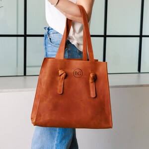 May include: A brown leather tote bag with two handles and a rectangular shape. The bag has a circular emblem with the letters "BG" on the front. The handles are attached with knots. The bag is held by a person wearing a white shirt and blue jeans.