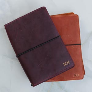 May include: Two leather journals, one in burgundy and the other in a rust colour, are shown. Each journal has a black elastic closure and gold initials. The burgundy journal has "NN" and the rust journal has "AG".