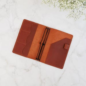 Könnte beinhalten: Ein Notizbuchumschlag aus hellbraunem Leder mit glatter Innenseite, drei vertikalen Kartenfächern auf der linken Seite, einer diagonalen Tasche auf der rechten Seite und drei schwarzen Gummibändern in der Mitte zur Befestigung von Einlagen.