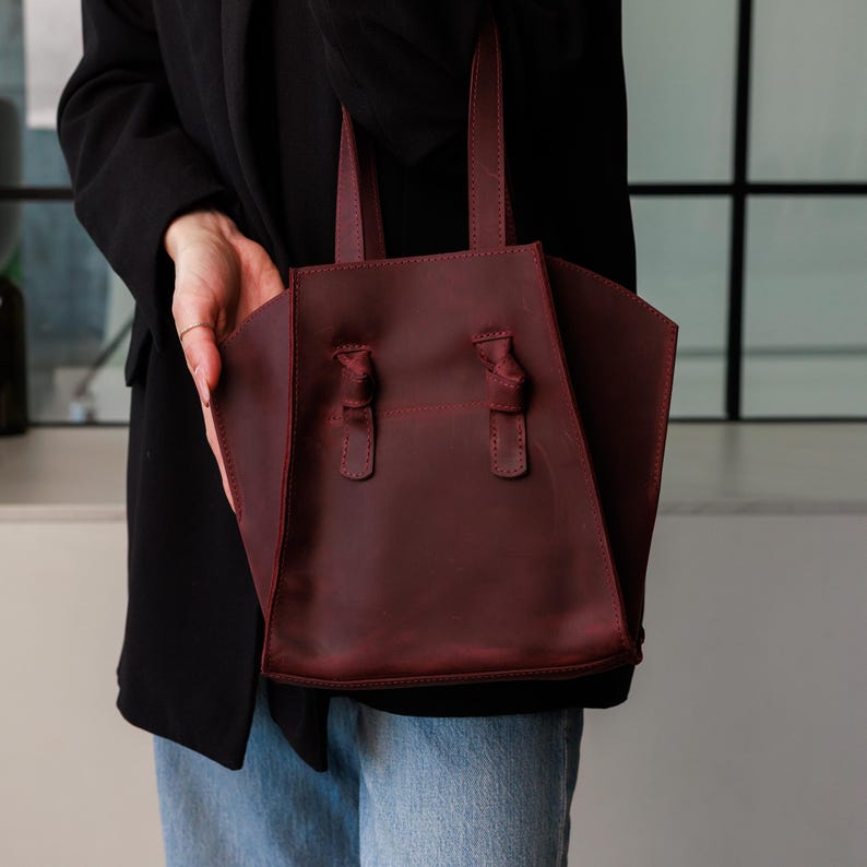 May include: A burgundy leather tote bag with knotted handle accents. The bag has a structured design with a rectangular base and a trapezoidal shape. The handles are attached with knots. The bag is held by a person.
