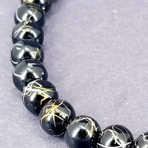 May include: A close-up of a black beaded bracelet. The beads are round and have a glossy finish. Each bead is decorated with a white and gold-colored, web-like pattern. The bracelet is strung on a black elastic cord.