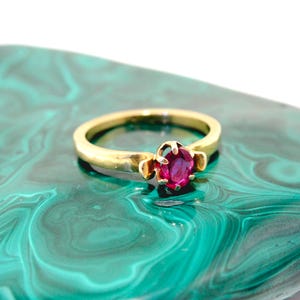 May include: A gold ring with a round, deep pink gemstone set in the center. The ring is displayed on a green and white marbled surface. The ring's band is a simple, polished gold.