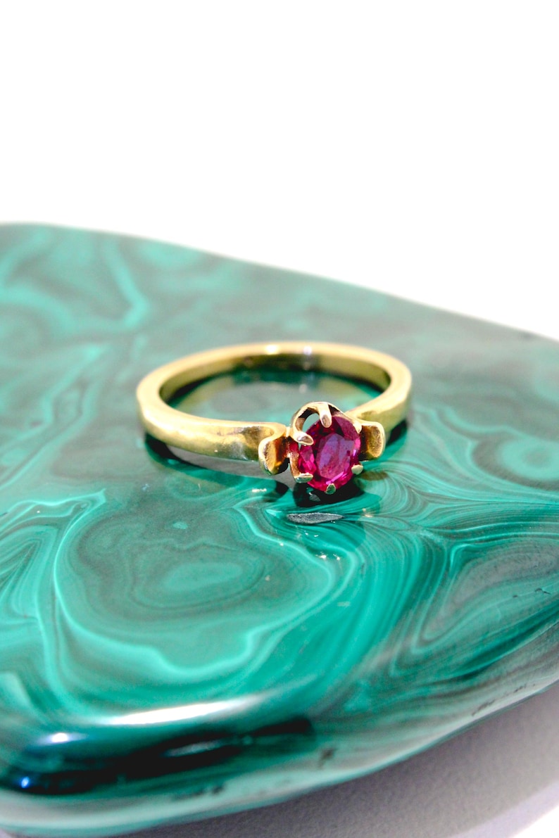 May include: A gold ring featuring a round, deep pink gemstone. The ring is set on a green and white marbled surface. The ring's band is a simple, polished gold. The gemstone is a vibrant focal point.