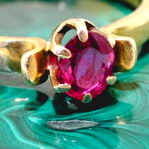 May include: A gold ring with a vivid, oval-cut ruby gemstone. The ruby is a deep pink-red hue, held in place by a traditional prong setting. The ring's band is polished gold, providing a strong contrast with the gemstone.
