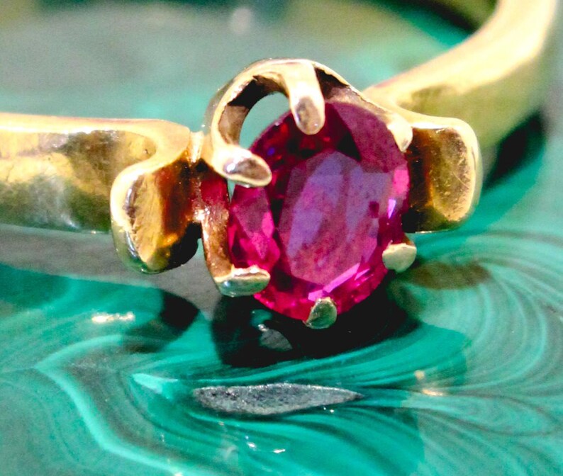 May include: A gold ring with a vivid, oval-cut ruby gemstone. The ruby is a deep pink-red hue, held in place by a traditional prong setting. The ring's band is polished gold, providing a strong contrast with the gemstone.