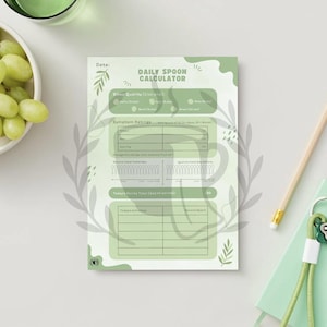 May include: A light green Daily Spoon Calculator notepad with a minimalist design, featuring sections for symptom ratings and daily notes. A pencil, a small green notebook, and a bunch of green grapes in a white bowl are also visible.
