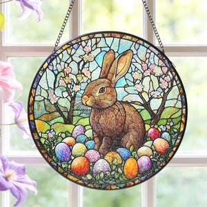 May include: Circular stained glass window art depicting a brown rabbit and Easter eggs. The design includes trees and flowers, set within a black metal frame and suspended by a chain. The artwork is a decorative piece.