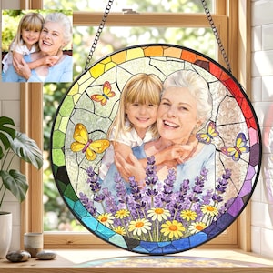 Puede incluir: Una vidriera redonda colgada de una cadena negra. Presenta una foto de una mujer y una niña, rodeadas de mariposas, lavanda y margaritas. El borde es un mosaico de vidrio de colores.