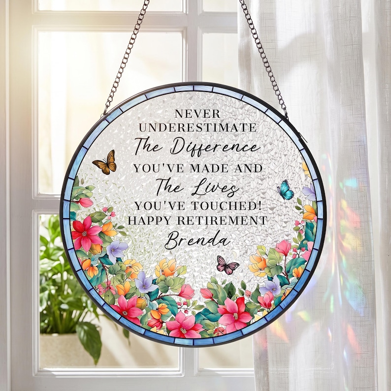 May include: Round stained glass window art with a floral design and butterflies. The text reads "NEVER UNDERESTIMATE The Difference YOU'VE MADE AND The Lives YOU'VE TOUCHED! HAPPY RETIREMENT Brenda". The window is framed in black and hangs from a chain.