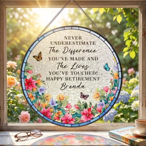 May include: A round, stained-glass window hanging with a black metal chain. The window features a floral design with butterflies and the text "NEVER UNDERESTIMATE The Difference YOU'VE MADE AND The Lives YOU'VE TOUCHED! HAPPY RETIREMENT Brenda."