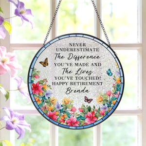 May include: Round stained glass window art with a black metal chain for hanging. The window features the text "NEVER UNDERESTIMATE The Difference YOU'VE MADE AND The Lives YOU'VE TOUCHED! HAPPY RETIREMENT Brenda" with floral and butterfly accents.