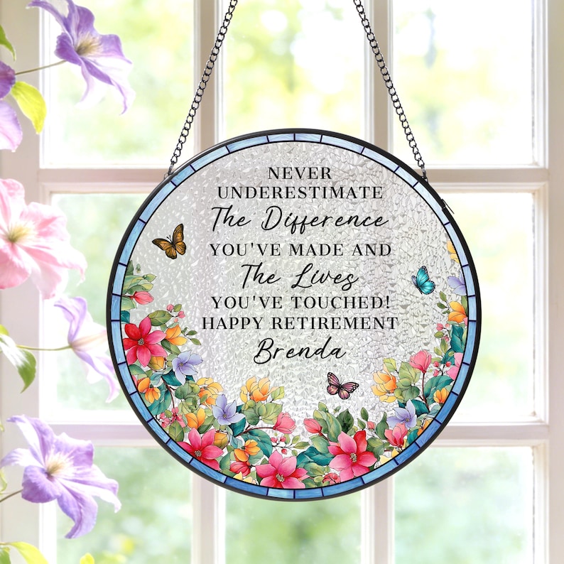 May include: Round stained glass window art with a black metal chain for hanging. The window features the text "NEVER UNDERESTIMATE The Difference YOU'VE MADE AND The Lives YOU'VE TOUCHED! HAPPY RETIREMENT Brenda" with floral and butterfly accents.