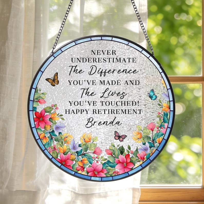 May include: A round, stained-glass window hanging with a floral border in shades of pink, yellow, and purple. The text reads "NEVER UNDERESTIMATE The Difference YOU'VE MADE AND The Lives YOU'VE TOUCHED! HAPPY RETIREMENT Brenda."