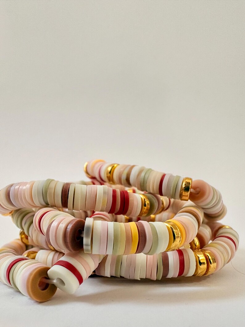 Peut inclure: Plusieurs bracelets empil&eacute;s compos&eacute;s de perles plates en forme de disque dans des tons pastel de rose, blanc, rouge, jaune et marron. Des entretoises en m&eacute;tal dor&eacute; sont intercal&eacute;es entre les perles. Les bracelets sont empil&eacute;s.