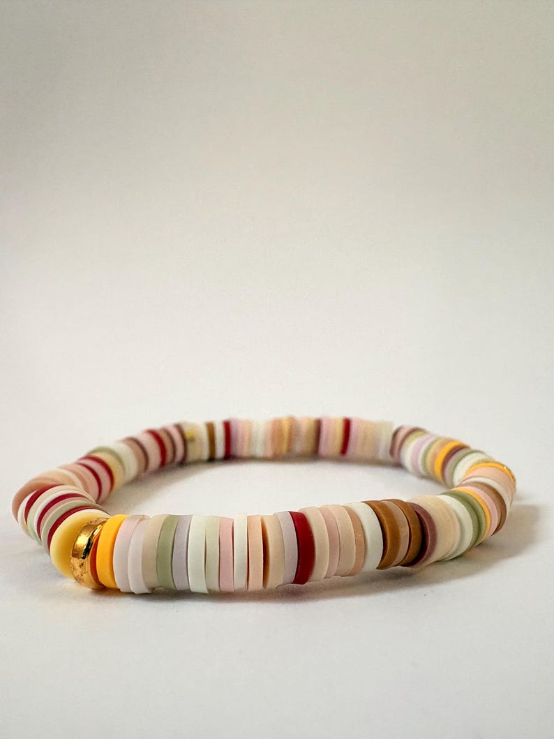 Peut inclure: Un bracelet de perles color&eacute;es avec des perles plates et rondes de diff&eacute;rentes couleurs, dont le rouge, le jaune, le blanc, le rose et le marron. Le bracelet a un fermoir dor&eacute;. Les perles sont enfil&eacute;es pour former une forme circulaire flexible.