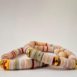 Peut inclure: Deux bracelets de perles aux couleurs pastel, rose, blanc, jaune et vert. Les bracelets sont orn&eacute;s d'&eacute;l&eacute;ments dor&eacute;s. Les perles plates et en forme de disque cr&eacute;ent un effet de superposition.