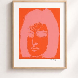 May include: Framed pop art print featuring a stylized face with pink hair and facial features against a vibrant orange background. The artwork is signed in the bottom right corner and encased in a light wood frame.