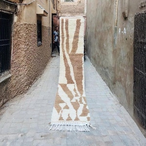 Op de afbeelding: Een lang, rechthoekig tapijt met een crèmekleurige achtergrond en bruine geometrische patronen. Het tapijt wordt verticaal weergegeven in een smalle steeg, waarbij het ontwerp en de textuur worden getoond. Het tapijt heeft een franje aan de onderkant.