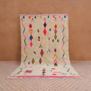 May include: Cream-colored, rectangular rug with a vibrant geometric pattern. The design features colorful diamond, zig-zag, and triangle shapes in pink, orange, blue, and green. The rug has a fringed edge and is displayed on a patterned tile floor.
