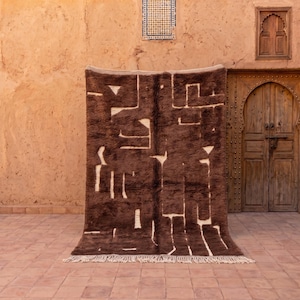 May include: A rectangular area rug featuring a deep brown, shaggy pile with a contrasting cream-colored geometric pattern. The design consists of irregular, angular lines and shapes throughout the surface, finished with a fringe along the bottom edge.