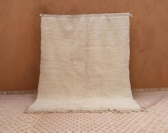 May include: A rectangular, off-white rug with a thick, textured pile. The rug has a fringe along the bottom edge and is displayed against a brown wall. The rug's neutral color and simple design make it suitable for various interior styles.