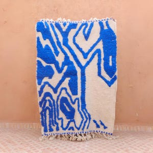 May include: A rectangular, off-white rug with a bold blue abstract design. The pattern resembles a stylized tree or network of veins. The rug has a fringe along the bottom edge and is placed on a pink tiled floor.