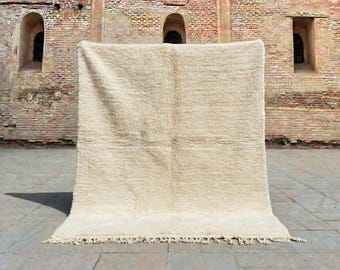 May include: A large, off-white rug with a textured surface and a fringe along the bottom edge. The rug is displayed against a brick wall with arched windows. The rug appears to be made of a thick, woven material.