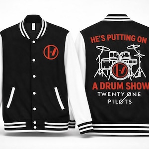 Peut inclure: Veste universitaire noire et blanche avec des manches blanches. Le devant présente un logo rouge, tandis que le dos affiche un dessin de batterie et le texte "HE'S PUTTING ON A DRUM SHOW TWENTY ONE PILOTS."