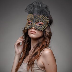 May include: A woman wearing a decorative mask with a black veil and a gold and black design. The mask covers the eyes and forehead. The woman has long, wavy brown hair and is wearing a beige top.