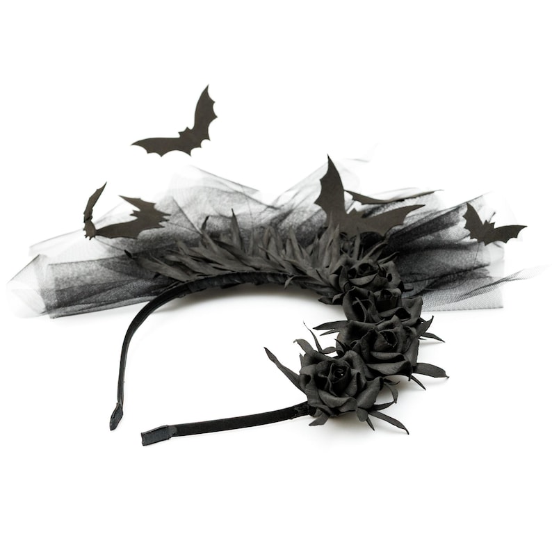 May include: A black headband with black roses, feathers, and tulle. The headband is decorated with several black paper bats, creating a gothic or Halloween theme. The design is set against a white background.