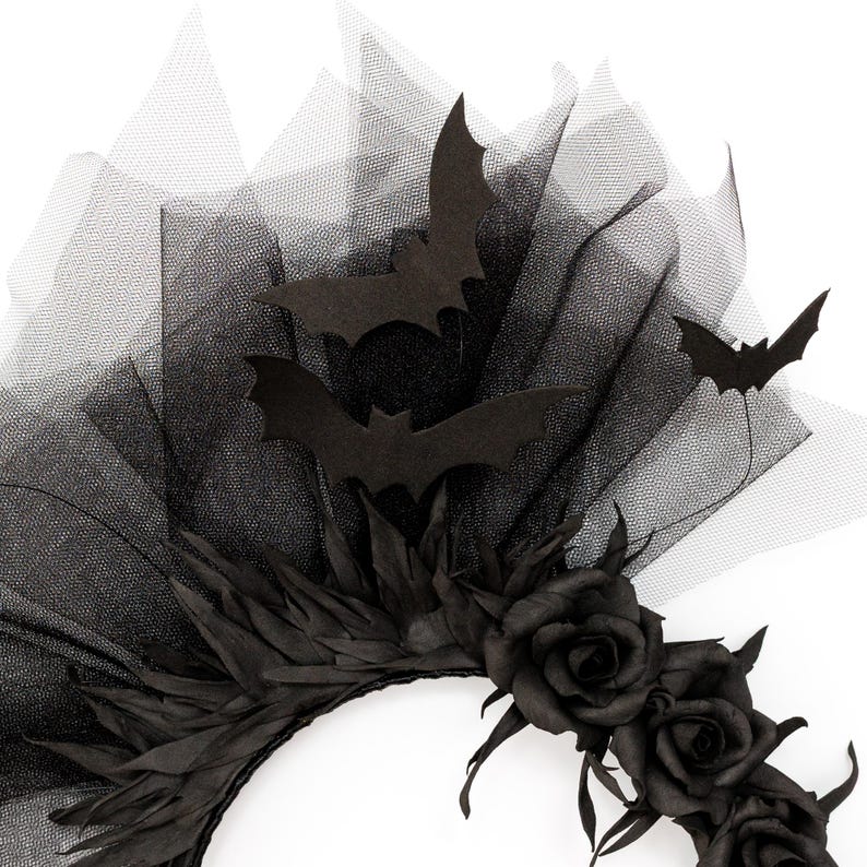 May include: A black Halloween headpiece featuring a black tulle veil, black paper bats, and black roses. The headpiece has a circular base with black leaves and three black roses. The bats are in various sizes and positions.