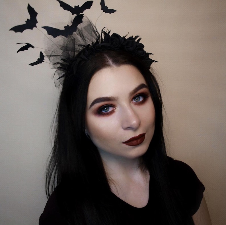 May include: A black Halloween headband with bat decorations. The headband features a black floral design and a sheer black veil. Several black bat cutouts are attached to the headband, creating a spooky and festive look. The person in the image is wearing dark makeup.