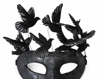 May include: A black masquerade mask with a textured, cracked surface finish. The top of the mask is adorned with several black bird figures in various flight positions, attached by thin wires, creating a layered, three-dimensional effect.