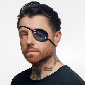 May include: Black leather eye patch with metal eyelets and a thin strap. The eye patch is worn over one eye. The person in the image has a beard and tattoos on their neck. The background is plain white.