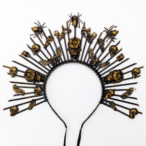 May include: A black and gold Halloween-themed headpiece with a halo design. The headpiece features a series of radiating spikes adorned with skulls, spiders, and other gothic-inspired embellishments. The central skull is the focal point.