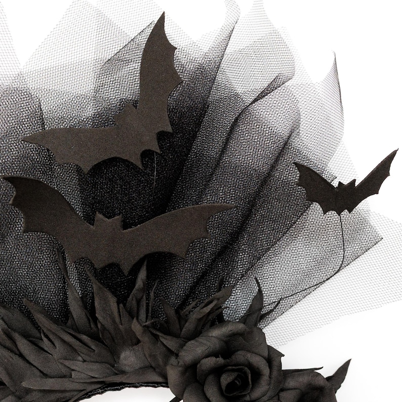 May include: A black Halloween-themed headpiece featuring felt bat cutouts, black tulle, and a black rose. The headpiece has a layered design with the bats seemingly flying above the tulle. The overall aesthetic is dark and gothic.